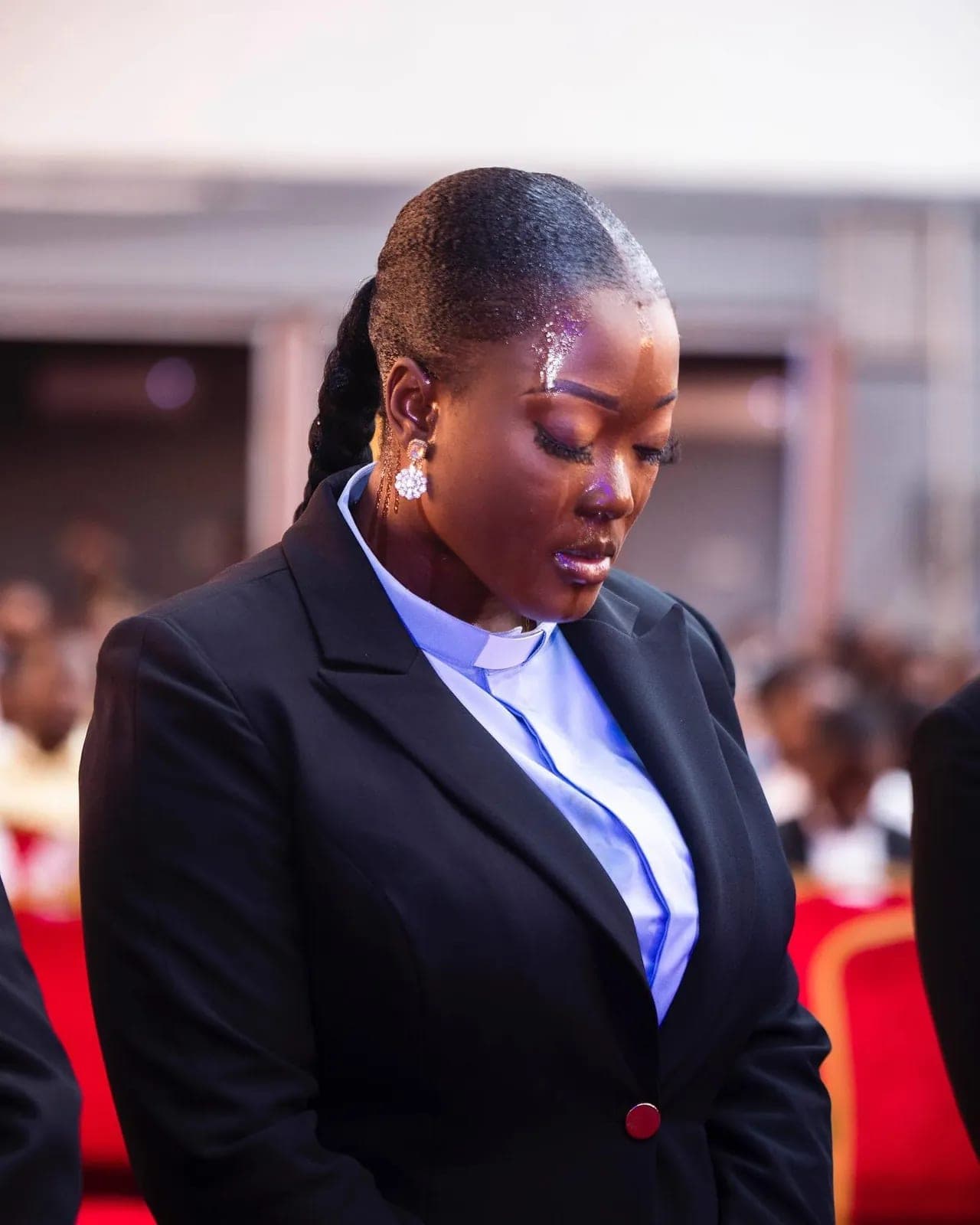 Petra Odubayo in a clerical collar and black suit, head bowed